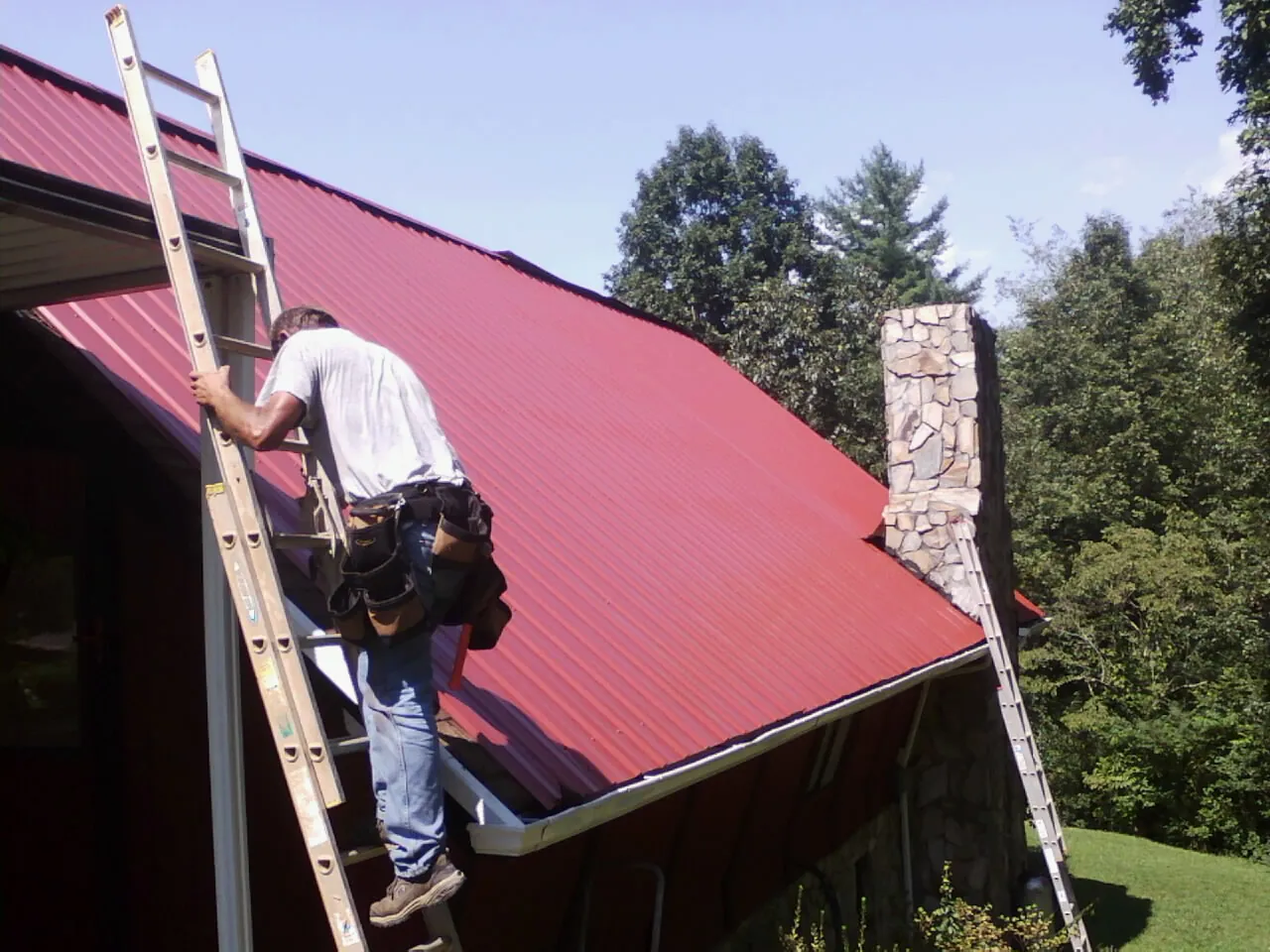 Roofer on a ladder beside a red metal roof and stone chimney — Soffit & Fascia Repair by Roofing Brothers in Mapleville